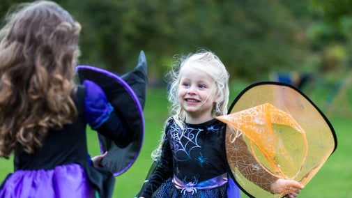 A young child dressed in a fancy dress witch costume for Halloween, holding an orange witches hat in her hand and smiling at another child also dressed in a fancy dress witch costume